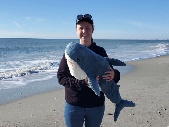 Sarah, a blond person in jeans, a black hoodie, and a ball cap. They’re holding a large blue and white plush shark, while standing on a peaceful sandy beach with the ocean in the background 