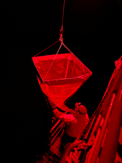 Large net being retrieved under red light on the side of a ship against a dark background.