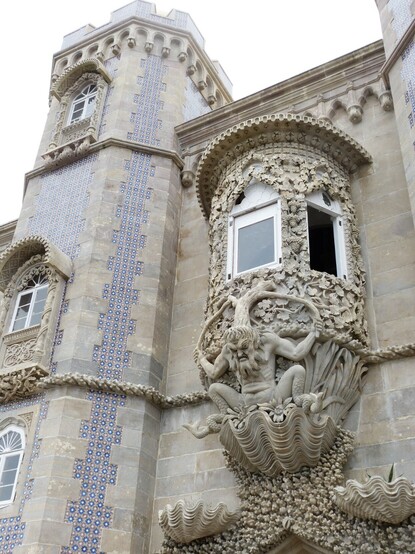 A carving of the mythological triton, with a tree growing from its head supporting a  bay-type window, with the triton standing on large clams and corals. The building housing the window has flat grey stones and a tower of flat grey stones with blue and white tile insets.