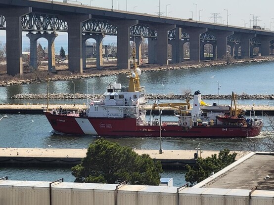 Red and white ship with Limnos, Canadian Coast Guard, and Fisheries and Oceans Canada on the side sailing through a canal with a large bridge in the background.