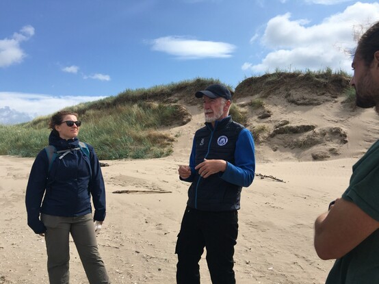 one person speaking with two others facing them on a beach. In the background is a sand dune, to the left of the speaker, the sand dune is stable and has grasses growing on the surface. to the right of the speaker the sand dune vegetation has slumped and lots of bare sand is visible.