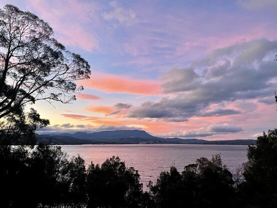 A landscape photo showing kunanyi / Mount Wellington in the distance, with water and lower hills in the foreground, framed by trees. The sky is still blue but with clouds coloured orange - yellow by the sunset. The orange colour is reflected in the water.