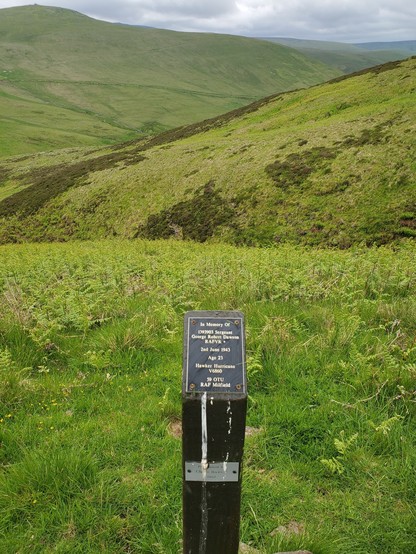A memorial post , a black plaque on a square wooden post, lies as grassy ground slopes steeply down to a hidden burn. Hillsides criss cross to a high ridge in the distnace under a grey sky.

This is the marker post for the crash of a Hawker Hurricane on 2 June 1943, killing its pilot Sgt G R Dawson RAFVR aged 23 years:

In Memory Of
1393903 Sergeant
George Robert Dawson
RAFVR +

2nd June 1943

Age 23

Hawker Hurricane V6860

59 OTU
RAF Milfield