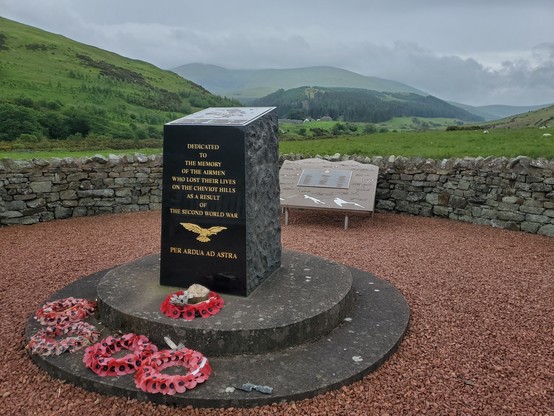 The Cheviot Memorial.

A black stone engraved in gold with the following stands on a round plinth, on which 5 poppy wreaths have been placed:

"Dedicated to the memory of the airmen who lost their lives on the Cheviot Hills as a result of the Second World War"

Then the emblem of the RAF

"Per Ardua Ad Astra"

on top of the stone is an engraved panel that lists all 19 crashes in the hills, and their details. The area enclosed by a dry stone wall and red chippings are on the ground. Behind, the beuatiful College valley, with green and wooded hills, sits under a cloudy sky