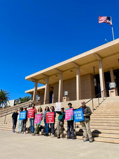 A group of people standing on the steps leading up to a modernist portico, holding signs with pro-science slogans 