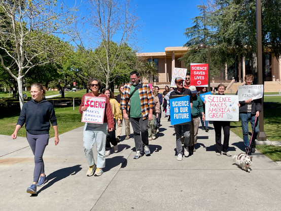 Same group same signs, marching in a group along a paved pathway through a green quadrangle in front of the building with the modernist portico 