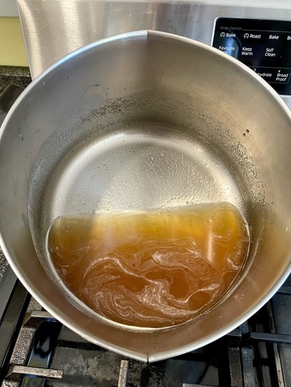 Looking down into a large cooking pot that is titled toward the camera to show the contents which is a little over a point of freshly boiled maple syrup. It is a fabulous caramel/amber color and still has tiny foamy bubbly steaks.