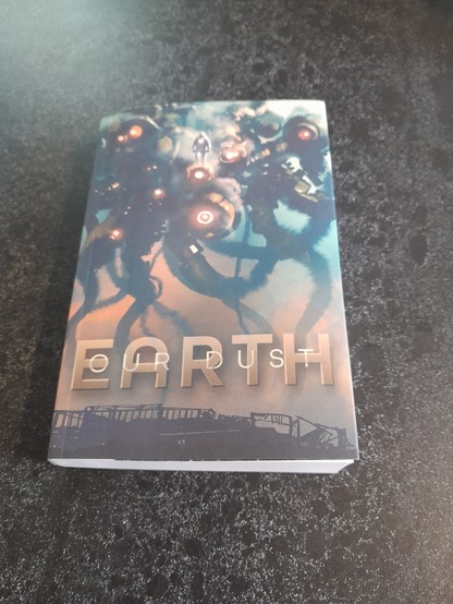 The book Our Dust Earth on a countertop.  Front cover is a ginormous biomech squid robot out for a post-apocalyptic stroll with his lil spacesuited buddy...