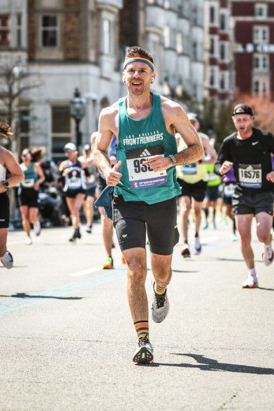 A man in short running tights, a tank top printed with the Los Angeles Frontrunners logo, and a rainbow headband, running in a race down a city street