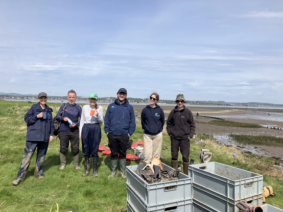 six smiling people standing behind a pile of grey boxes (now empty of plants) and in front of newly planted saltmarsh. 