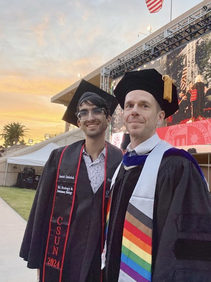 A man in graduation robes and a man in doctoral robes in front of a building and outdoor stage, under a sunset-lit sky