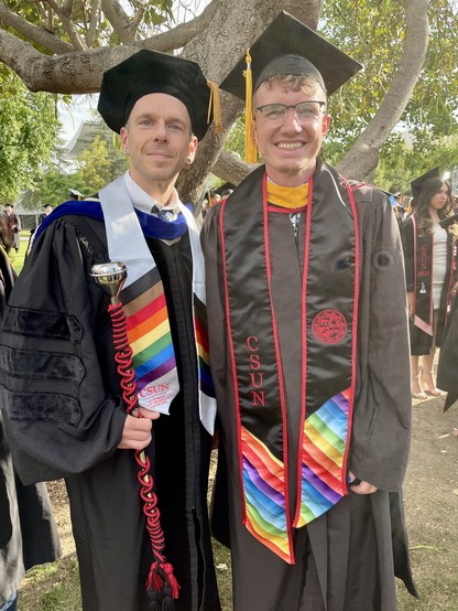 Two men, one in doctoral robes and one in Master's robes, standing under a green tree