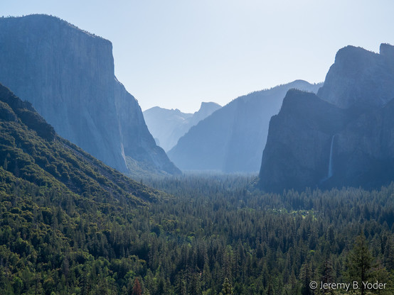 A green-forested valley with a flat floor, running into blue misty distance between steep vertical cliffs; on the right a waterfall is visible down one of the cliffs, and the distinctive profile of Half Dome is visible at the far end of the valley