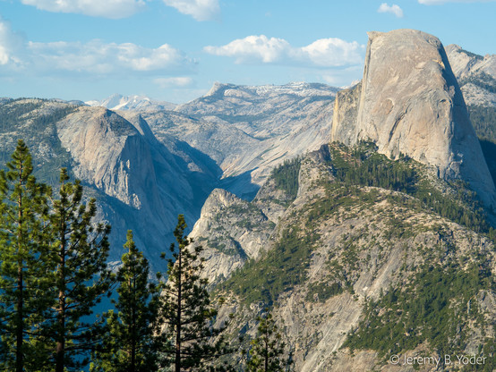 A view from the Glacier Point lookout, looking over pine trees to the eastern end of the Yosemite Valley; the valley in shadow to the right of Half Dome, a prominence of bare rock viewed edge-on with a sheer flat face on the left, and a rounded back on the right; all under pale blue sky studded with clouds
