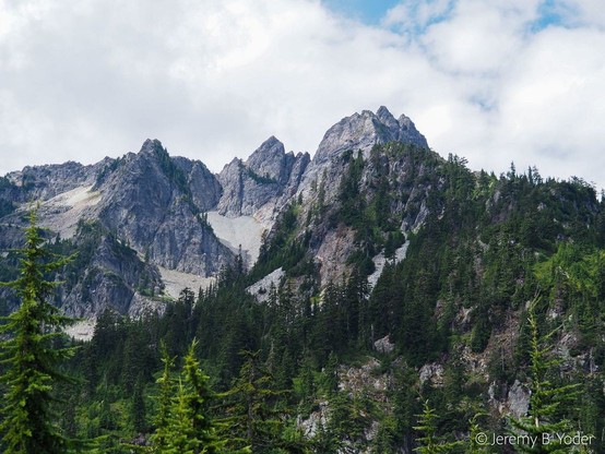 A rocky ridgeline with multiple sharp peaks, the lower slopes forested with green conifers, under white clouds with a little blue sky peeking through