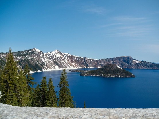 A deep blue arc of quiet lake, ringed by snow-streaked crater walls and a handful of green conifers, with a conical island rising from the water