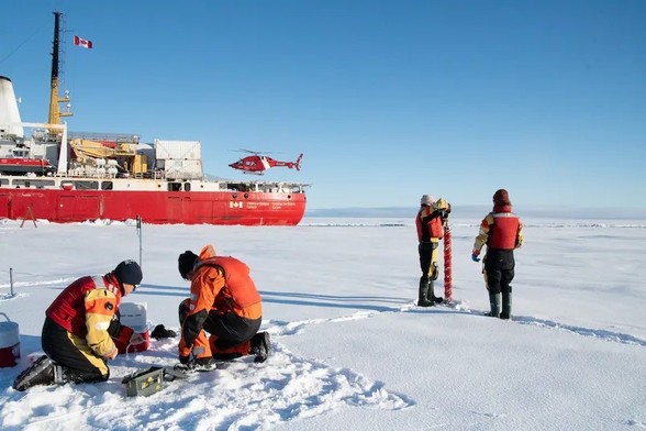 image/jpeg researchers in orange life suits take measurements on a snow covered ice sheet that extends into the distance. A red ship with a helicopter and Canadian flag sits in the background. Source: Amundsen Science.